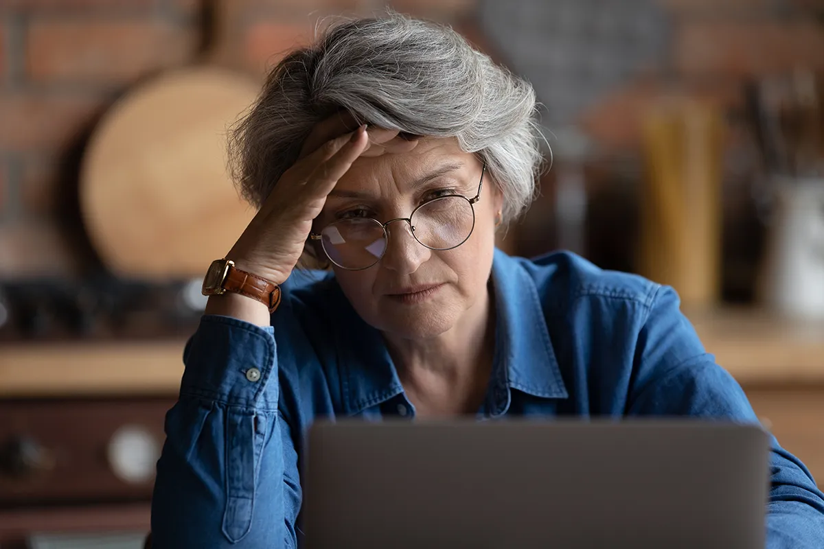 An elderly woman holds a hand to her head while staring at a computer screen